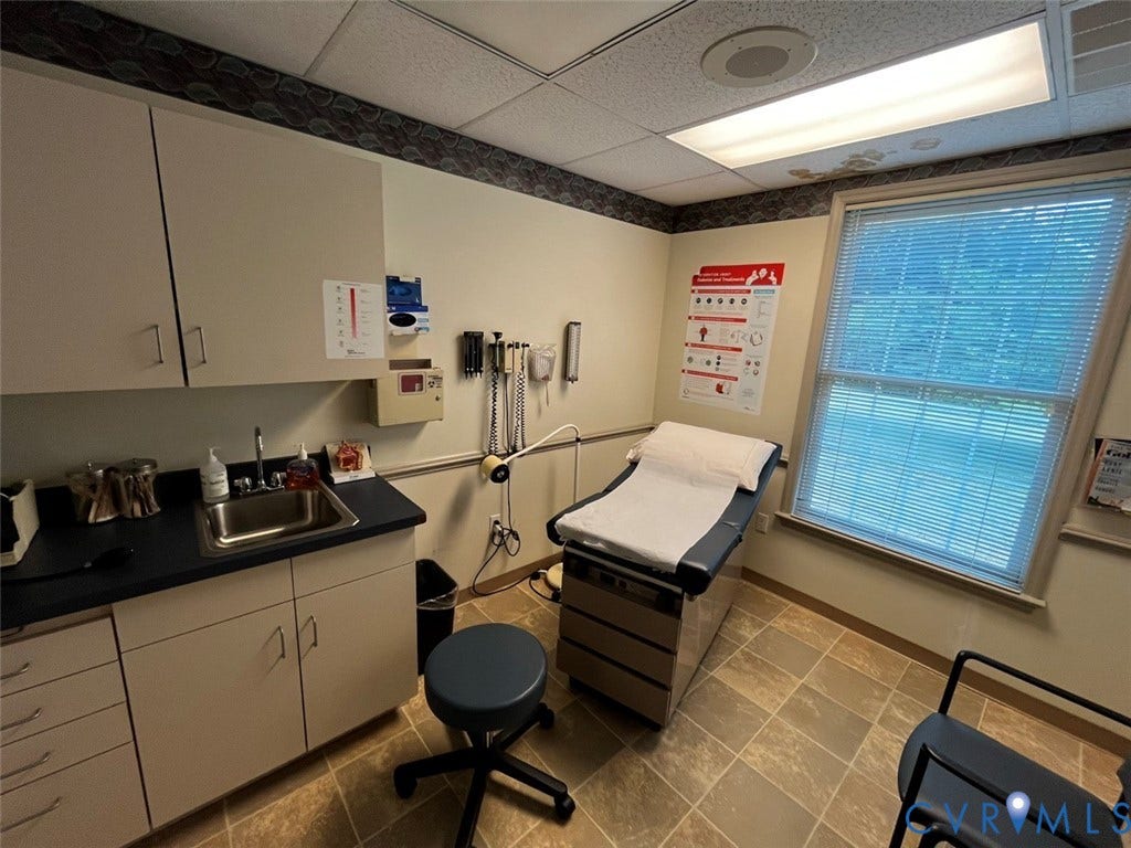 4064 James Madison Highway, Fork Union, VA 23055 Examination room featuring a built-in sink cabinet with overhead storage, a large window with horizontal blinds, and patterned floor tiling