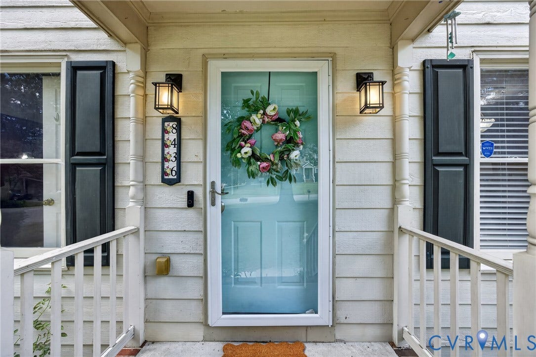 8436 Sir Lionel Place, Kingsland Wood, Ampthill, VA 23237 Light blue entry door with a white storm door, flanked by two windows with dark shutters, and a white porch railing