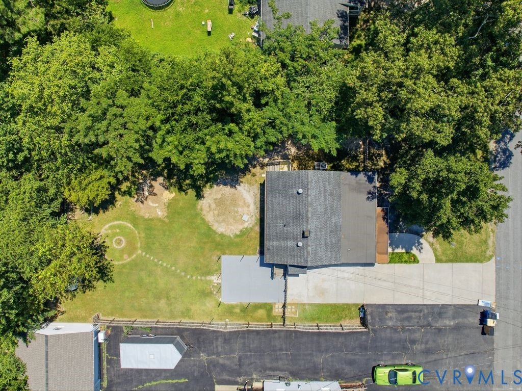 Aerial view highlighting a dark shingle roof, a light gray patio, and extensive surrounding mature trees