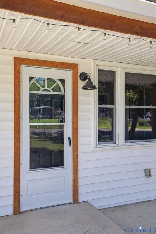 White siding exterior featuring a glass-paneled entry door with wood-finish trim, a barn-style outdoor light fixture, and string lights on a wooden beam