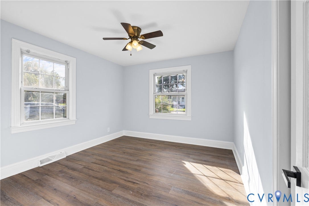 3516 Virginia Street, Woodlawn, Hopewell, VA 23860 Light-filled room featuring wood-finish flooring, two large windows, a ceiling fan with integrated lighting, and a white door with matte black hardware
