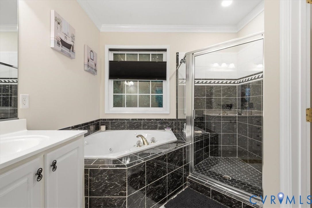 Corner jetted tub with tile surround, glass-enclosed shower with tiled walls and hexagonal floor, single vanity with white cabinet, and a window with blinds