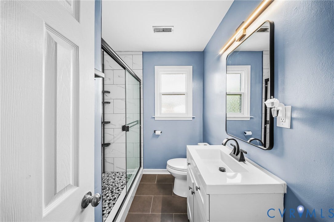 3304 Carlton Road, Henrico, VA 23223 Modern bathroom featuring a single white vanity with an integrated sink and matte black faucet, a rectangular illuminated mirror, a glass-enclosed shower with white subway tile and pebble flooring, and a white framed window