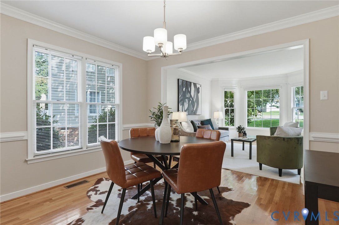 Dining area with hardwood flooring, a contemporary chandelier, crown molding, and trim, featuring a framed opening to a sunlit living space with a bay window