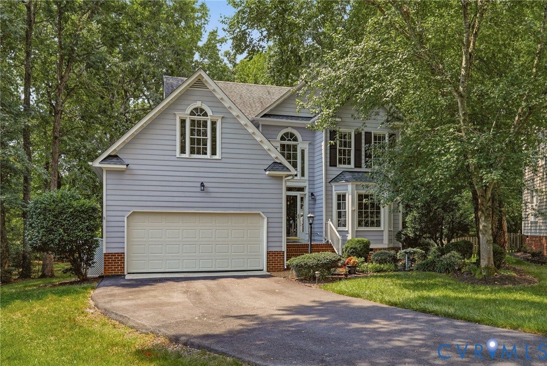 Light blue siding exterior featuring a two-car garage with a paneled door, an arched window dormer, multiple bay windows with dark shutters, and a landscaped front yard with mature trees