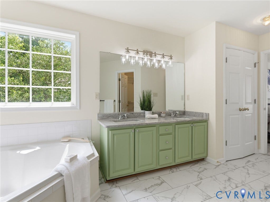 5707 Stephens Point Court, Chesterfield, VA 23831 Primary Bath w/ double windows featuring a spacious corner soaking tub, a dual vanity with a light green cabinet, a stone countertop, and a large mirror with a multi-bulb light fixture