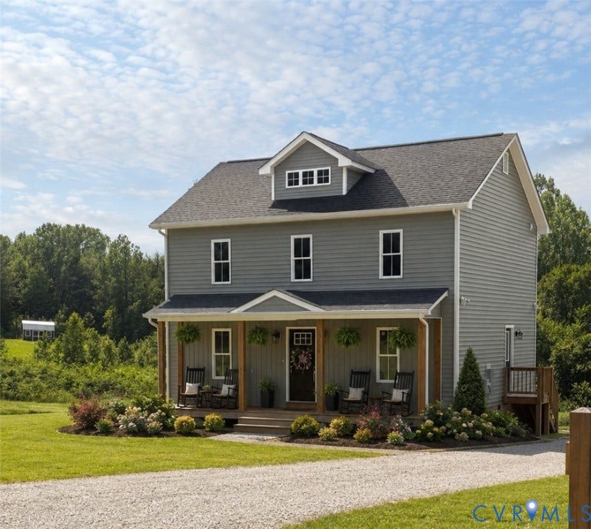 Two-story residence featuring grey horizontal siding, a covered front porch with wood columns, a dormer with multiple windows, and a gravel driveway Virtually Staged