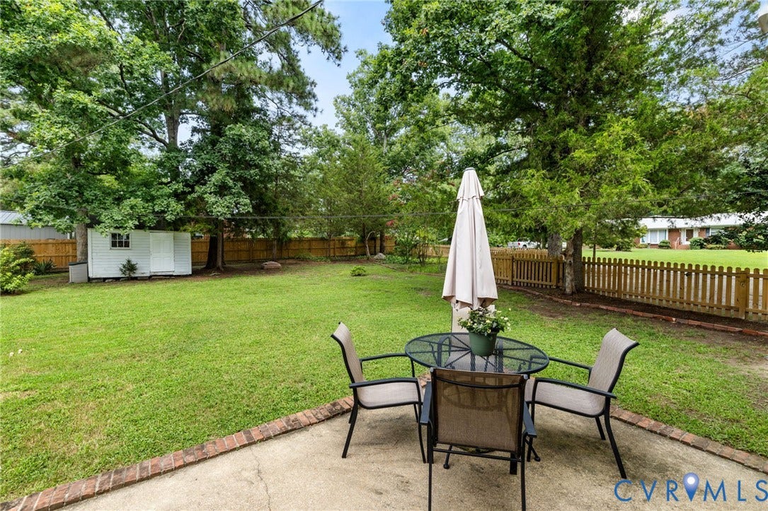 404 Aubery Road, Henrico, VA 23229 Expansive backyard featuring a concrete patio with brick edging, a manicured lawn, perimeter fencing, mature trees, and a white storage shed