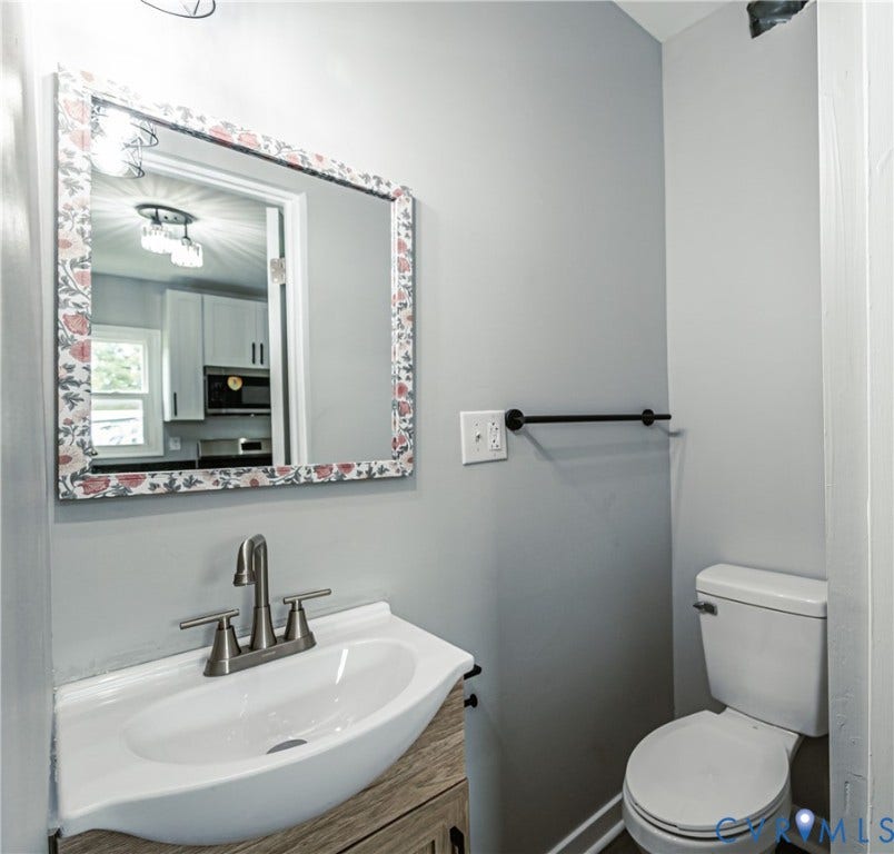 3903 Delmont Street, Henrico, VA 23222 Bathroom featuring a pedestal sink with a wood-finish vanity, a wall-mounted mirror with a decorative frame, a toilet, a black wall-mounted towel bar, and light gray wall paint