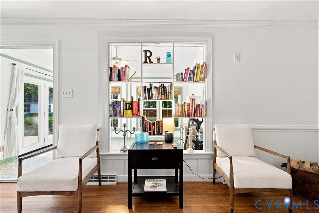 10204 Steuben Drive, Henrico, VA 23060 Bright and sophisticated living room showcasing hardwood flooring, paired lounge chairs with wood-frame construction, and a striking built-in bookcase wall with glass-front cabinetry displaying curated collections