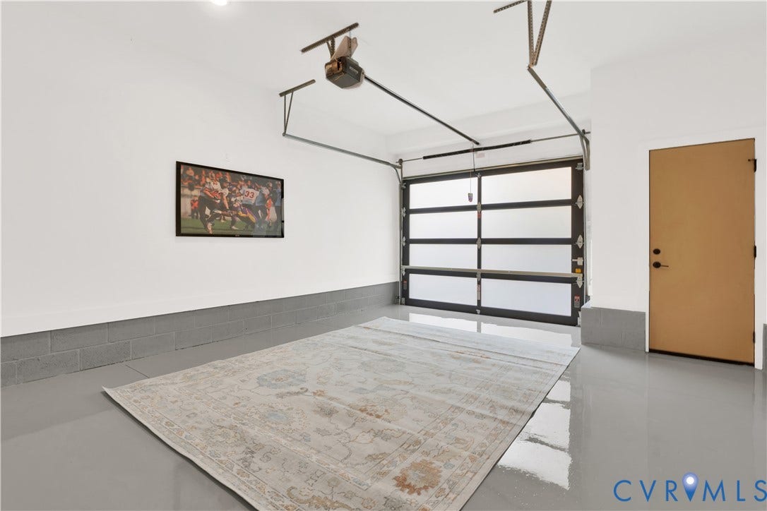 4506 1/2 Grove Avenue, Munford On Grove, Richmond, VA 23221 Spacious garage with a modern glass panel garage door, smooth gray epoxy flooring, white painted walls, and a light-colored interior door with matte black hardware