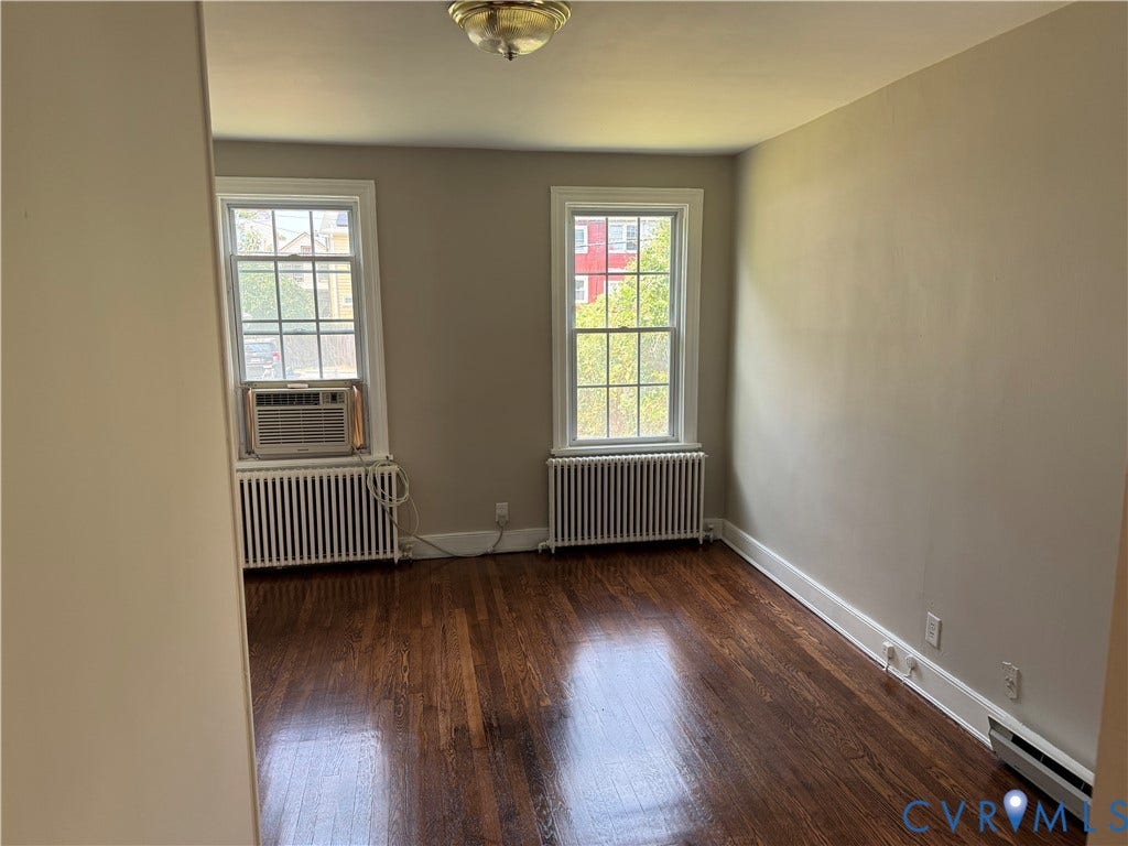 3118 Barton Avenue, Brookland Park, Richmond, VA 23222 Bright, light-filled room showcasing gleaming wood-finish flooring, dual tall windows with gridded panes, classic radiator heating, and neutral painted walls with crisp white trim