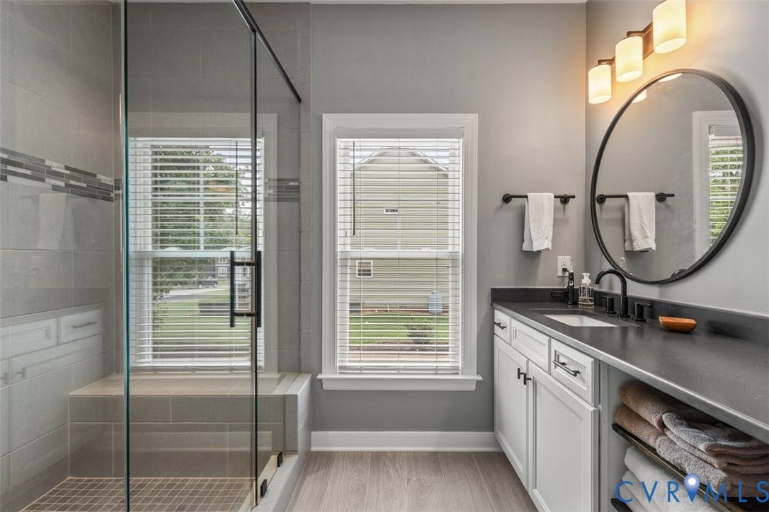 9081 Deerlake Drive, Charming, New Kent, VA 23124 Bathroom featuring a glass-enclosed shower with a built-in bench, a single vanity with dark countertop and white cabinetry, a round black-framed mirror, and wood-finish flooring
