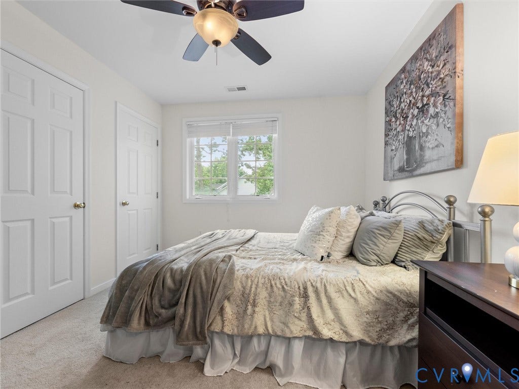 6100 Lilting Branch Way, Moseley, VA 23120 Serene primary bedroom featuring a metal bed frame, dual closets with six-panel doors, ceiling fan with integrated lighting, carpet flooring, and natural light from a multi-pane window