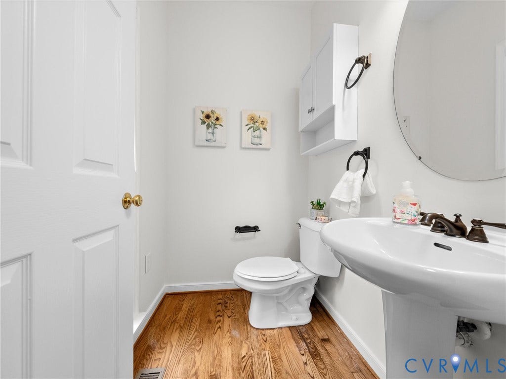 6100 Lilting Branch Way, Moseley, VA 23120 Wood-finish flooring, white pedestal sink, circular wall mirror, white toilet, and white wall-mounted cabinet