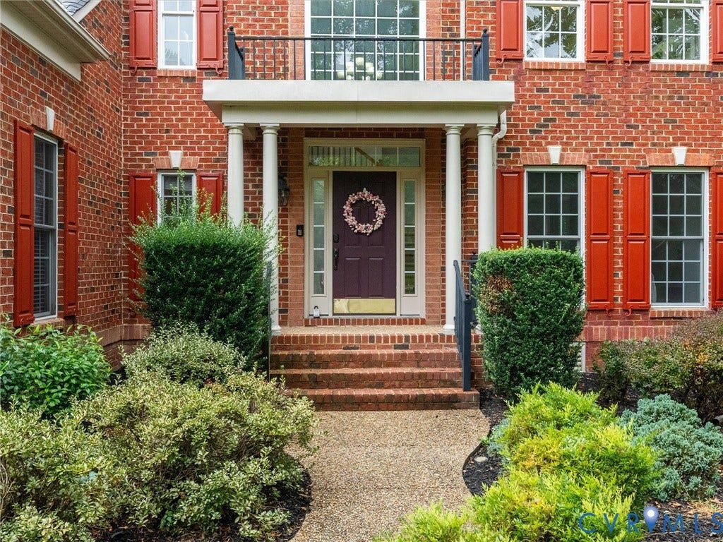 6100 Lilting Branch Way, Moseley, VA 23120 Brick facade with red shutters, portico with white columns, second-story balcony with black railing, and mature landscaping