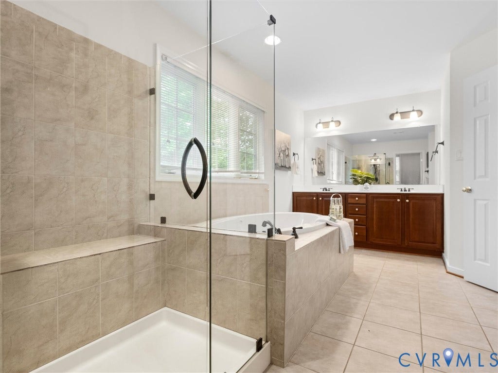6100 Lilting Branch Way, Moseley, VA 23120 Spa-inspired primary bathroom featuring a frameless glass-enclosed shower with tile surround, a soaking tub positioned for relaxation, dual vanities with wood cabinetry and ample storage, polished nickel fixtures, and neutral tile flooring throughout