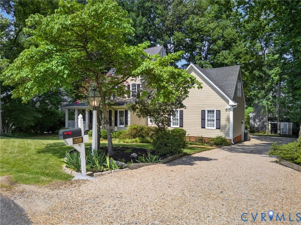 Light-colored siding exterior with dark shutters, gabled roofline, front porch with white columns, gravel driveway, and mature landscaping
