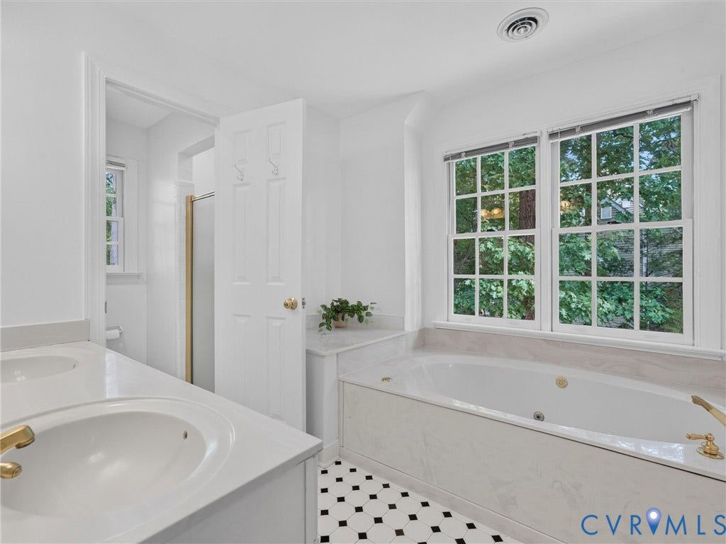 Dual vanity with undermount sinks and gold-finish fixtures, a large soaking tub with a tiled surround, checkerboard tile flooring, and multi-pane windows