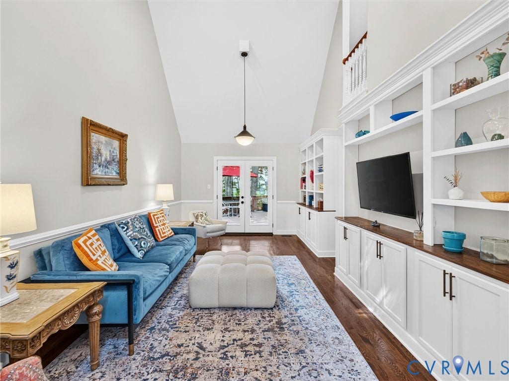 Vaulted ceilings with a pendant light fixture, extensive built-in shelving with lower cabinetry, rich wood-finish flooring, and French doors leading to exterior space