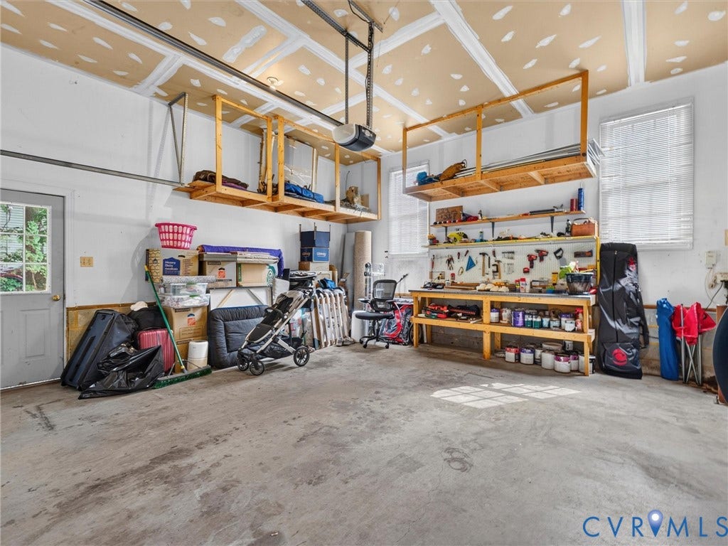 Garage featuring concrete flooring, built-in overhead storage shelves, a workbench with shelving, and multiple windows