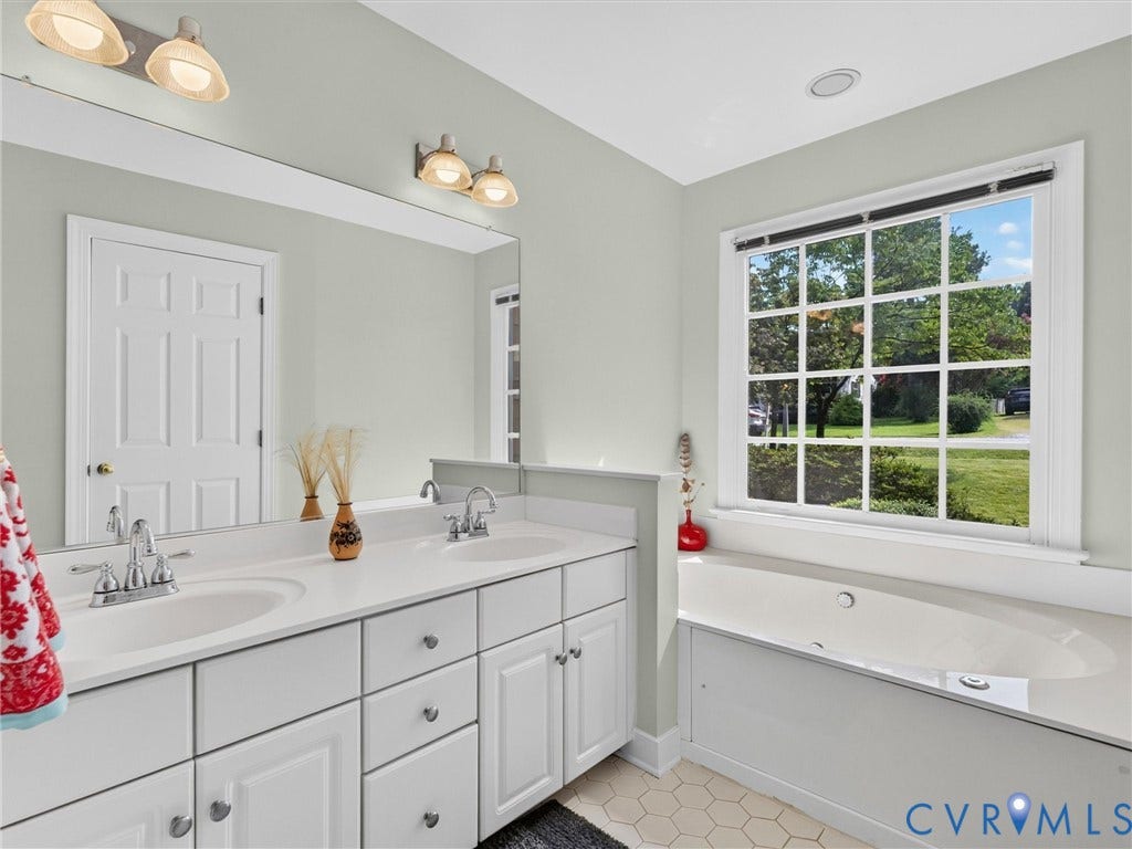 Double vanity with undermount sinks and chrome faucets, large mirror, built-in bathtub with tiled surround, hexagonal floor tiling, and a multi-pane window