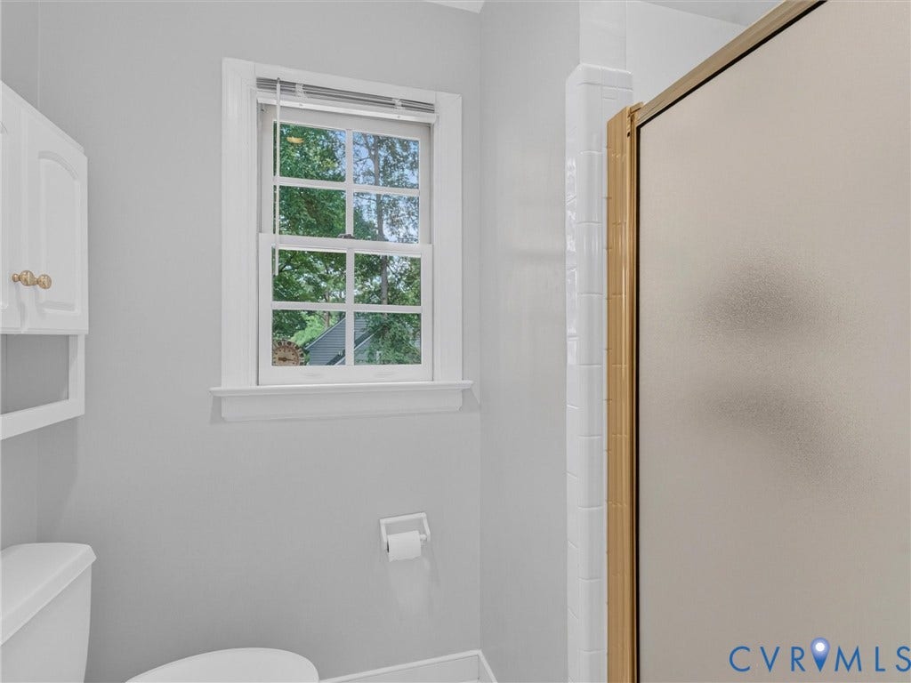 White framed window with privacy blinds, white wall-mounted cabinet, toilet, and a frosted glass shower enclosure