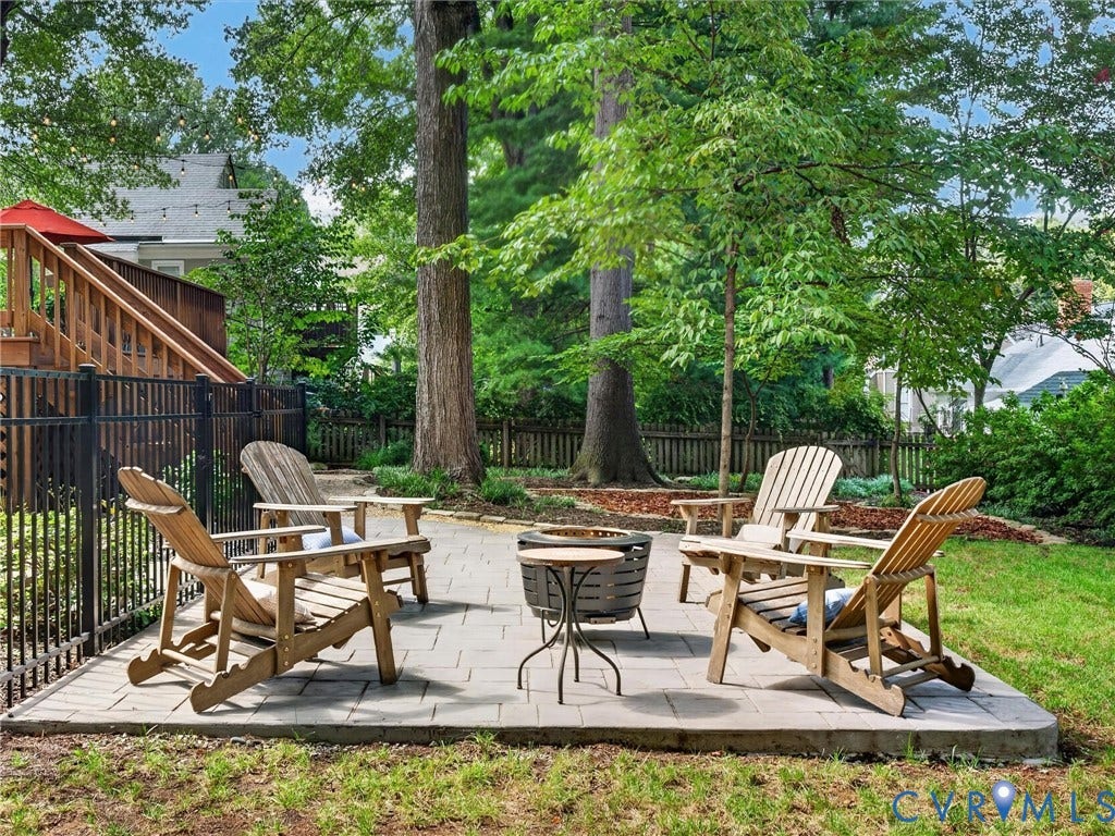 Paver patio with a fire pit, mature trees, wood deck, and black metal fencing