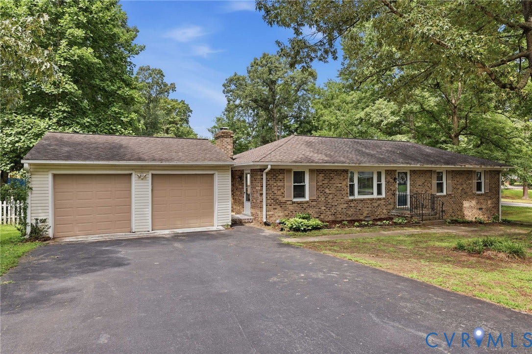3300 Woodsong Drive, Midlothian, VA 23112 Brick ranch-style residence featuring an attached two-car garage with tan doors, a paved driveway, and mature perimeter trees
