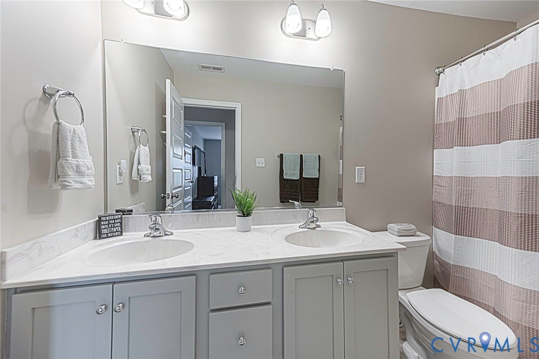 7205 Desert Candle Drive, Moseley, VA 23120 Guest bathroom featuring a dual vanity with light gray cabinetry and integrated sinks, a large wall-mounted mirror, overhead vanity lighting, and a toilet