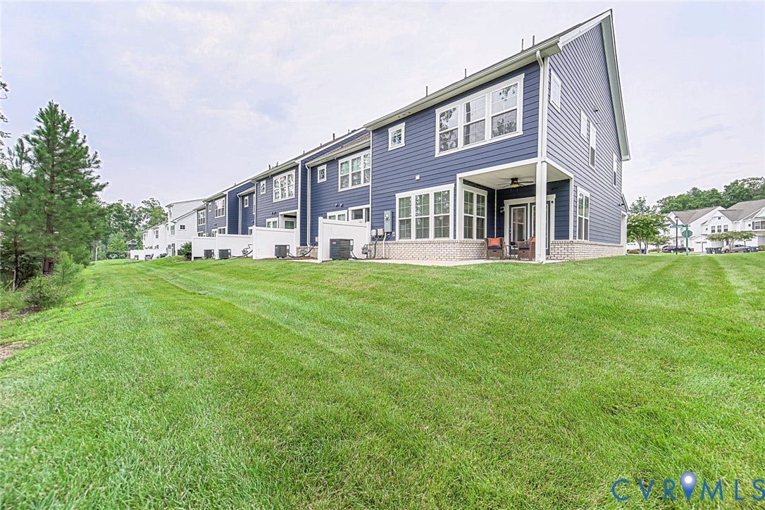 7205 Desert Candle Drive, Moseley, VA 23120 Rear exterior featuring blue horizontal siding, white trim, a covered patio with a ceiling fan, a brick foundation, and a large expanse of green lawn