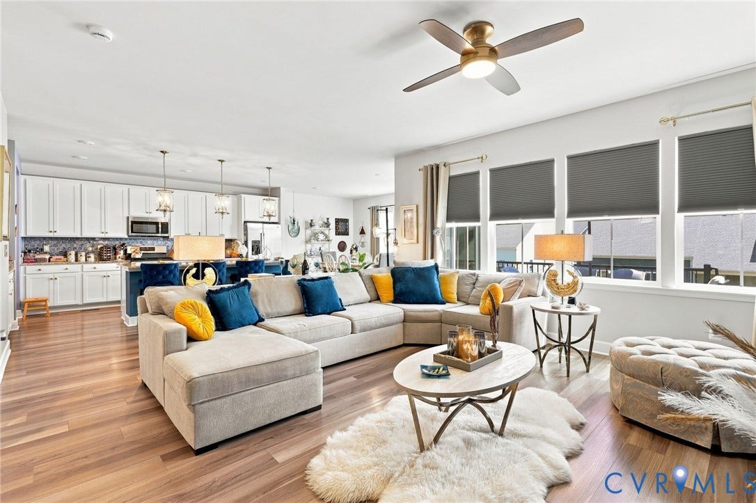 Open-concept living area featuring a light gray upholstered sectional sofa with chaise, hardwood flooring throughout, and abundant natural light from floor-to-ceiling windows with motorized roller shades