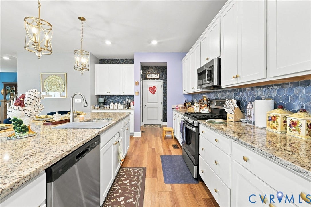 Galley kitchen featuring white cabinetry with brass hardware, granite countertops, stainless steel appliances including a black range, undermount sink with pull-down faucet, blue hexagonal tile backsplash, recessed and pendant lighting, and wood-finish flooring throughout