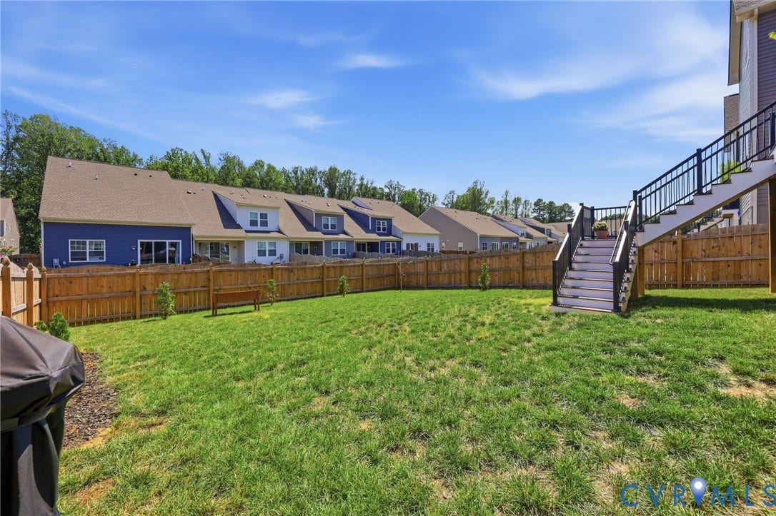 Expansive backyard featuring a manicured lawn with mature trees lining the perimeter, wooden privacy fencing, and a multi-story residence with distinctive architectural details including dormer windows and a covered deck with metal railings