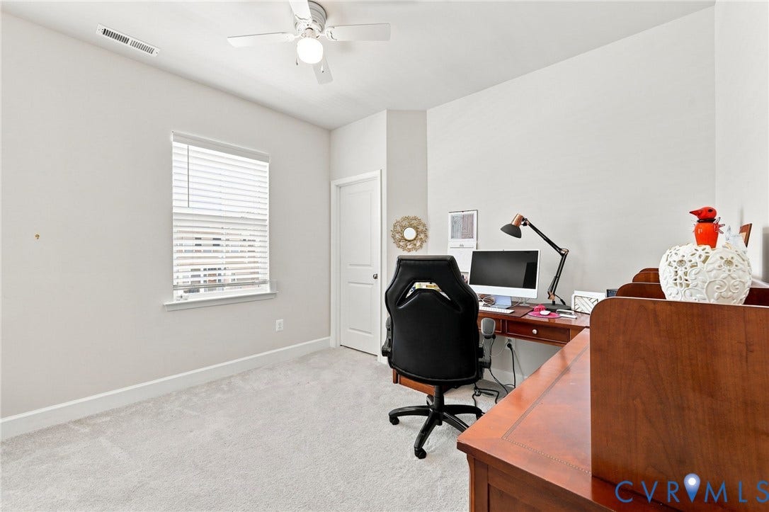 Bright home office featuring a spacious layout with neutral wall finishes, carpet flooring, a ceiling fan with integrated lighting, and large window with blinds providing natural light