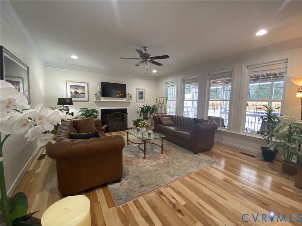 3267 Bayberry Lane, Goochland, VA 23063 Spacious living area featuring wood-finish flooring, a white mantel fireplace with an ornate screen, and triple windows providing natural light