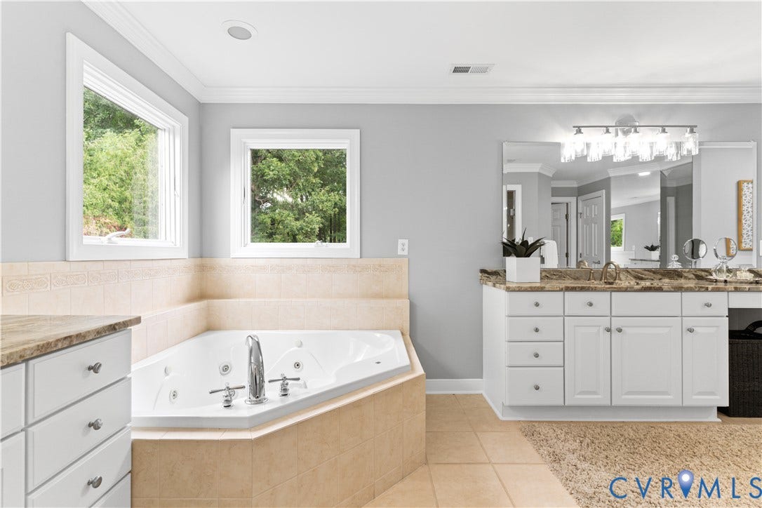 Corner jetted bathtub with a tiled surround, dual windows, and two separate vanity areas featuring stone countertops and integrated sinks
