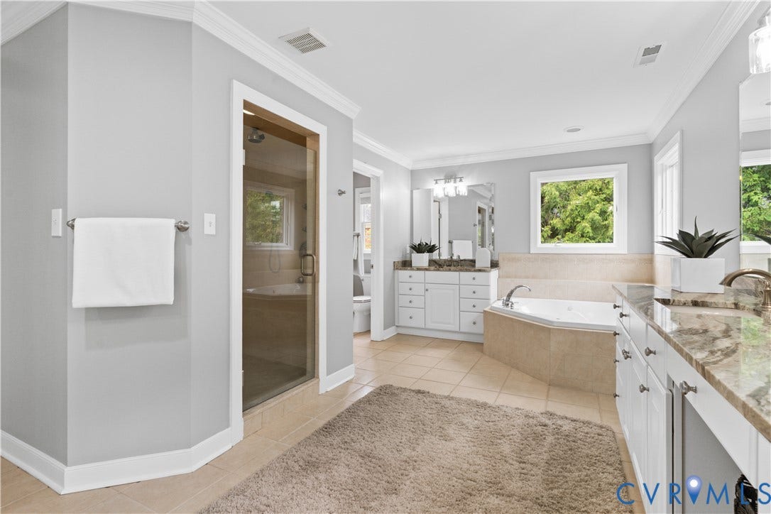 Spacious, updated primary bathroom featuring a glass-enclosed shower, corner soaking tub, and dual vanity areas with stone-finish countertops