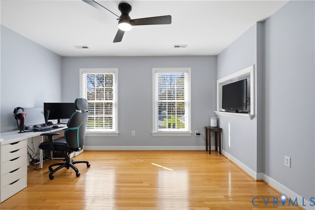 Hardwood-finish flooring throughout, complemented by two windows with horizontal blinds, a ceiling fan with integrated lighting, and a built-in media niche