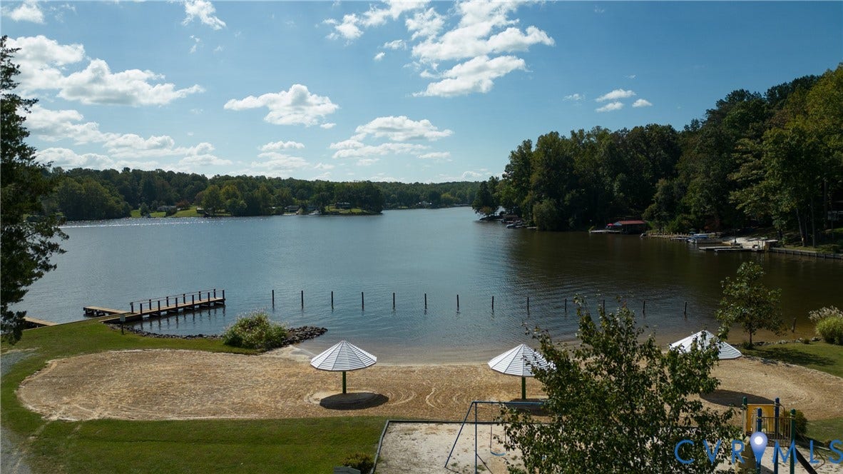 309 Ellis Drive, Louisa, VA 23093 Expansive lakefront property featuring a private sandy beach area, two sun shades, a wooden dock, and mature trees bordering the shoreline