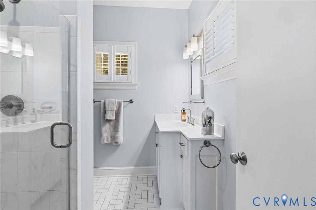 Bathroom featuring a glass-enclosed shower with white tiled walls, a single vanity with a white countertop and undermount sink, a wall-mounted mirror, and white plantation shutters on the window