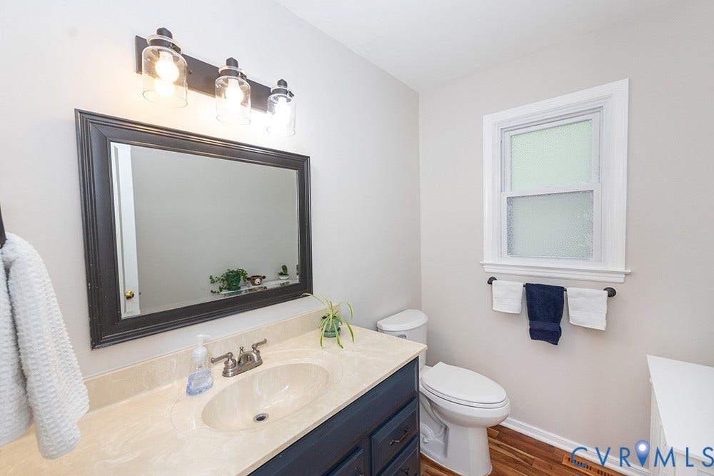 419 Rossmere Drive, Walton Park, Midlothian, VA 23114 Wood-finish flooring complementing a white toilet, dark blue vanity with a light countertop and integrated sink, and a framed mirror under a three-light fixture