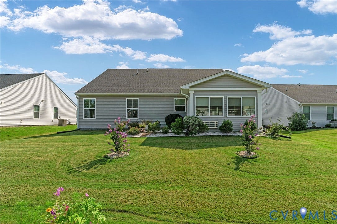 7484 Southamptonshire Way, Autumn Pines, New Kent, VA 23124 Expansive rear lawn featuring a sunroom addition with multiple windows, light gray siding, and a landscaped garden bed