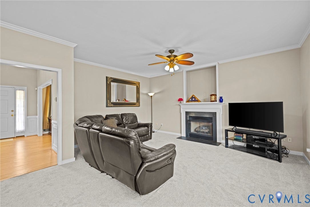 Carpeted living area featuring a white mantel fireplace with a black surround, ceiling fan with integrated lighting, crown molding, and a wide entryway to a foyer with wood-finish flooring