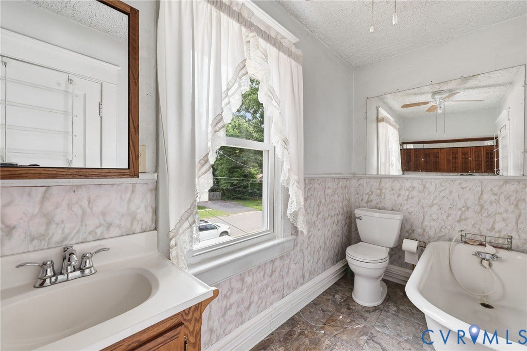 Bathroom featuring a wood-finish vanity with an integrated sink, a rectangular framed mirror, patterned wall paneling, a full bathtub, and a window with white curtains