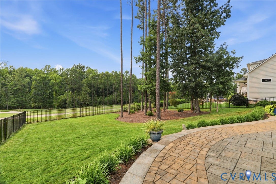 Expansive backyard featuring a patterned paver patio, a lush lawn, mature trees, and a black iron fence bordering a wooded area
