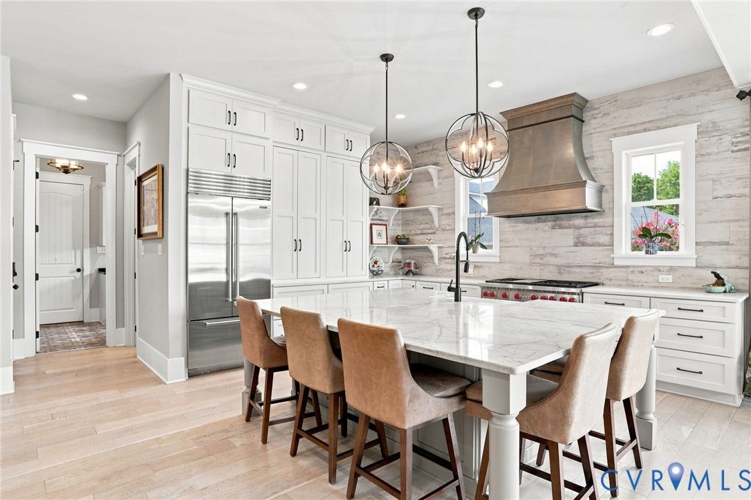Gourmet kitchen featuring a large central island with a stone countertop, white cabinetry with dark hardware, a professional-grade stainless steel range with a custom wood-finished hood, and light wood-finish flooring
