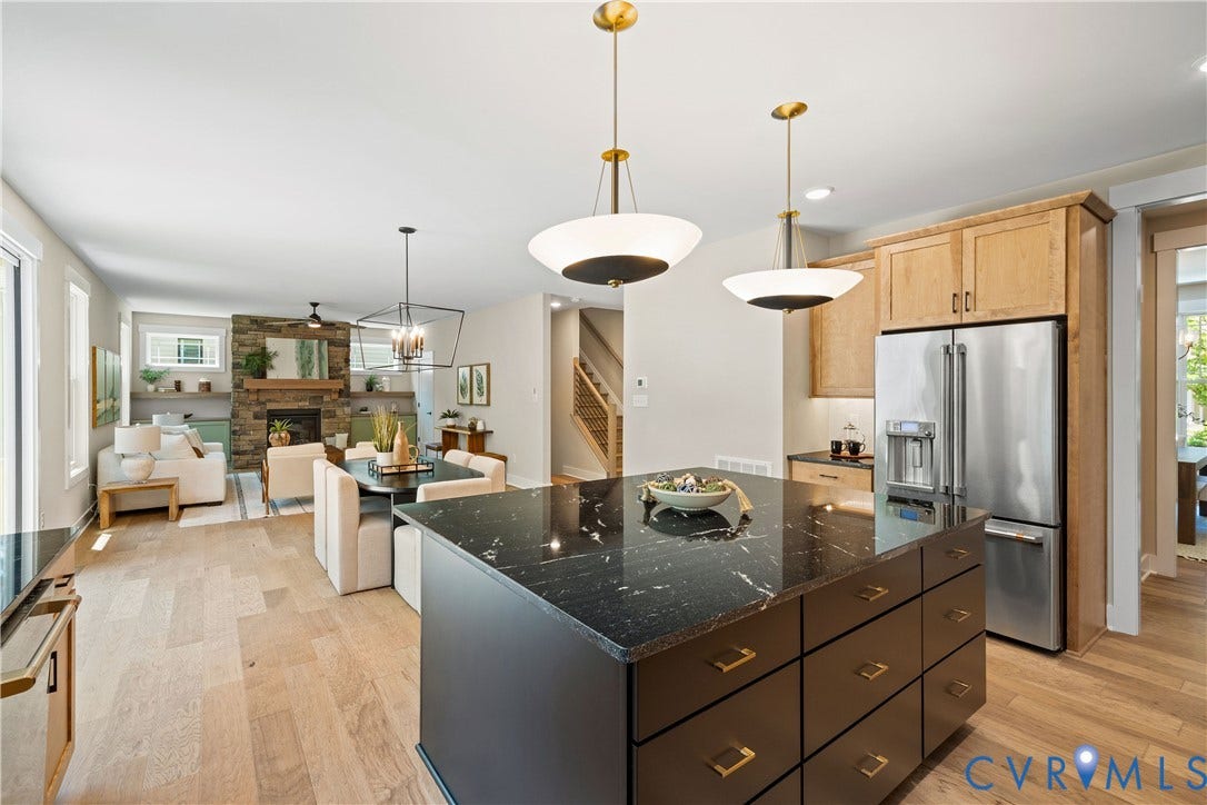 Open-concept living area featuring a kitchen island with dark stone countertop and brass hardware, light wood-finish flooring, a stacked stone fireplace, and stainless steel appliances
