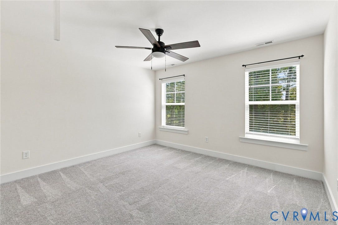 Light gray carpeting, two windows with white blinds, and a dark-toned ceiling fan