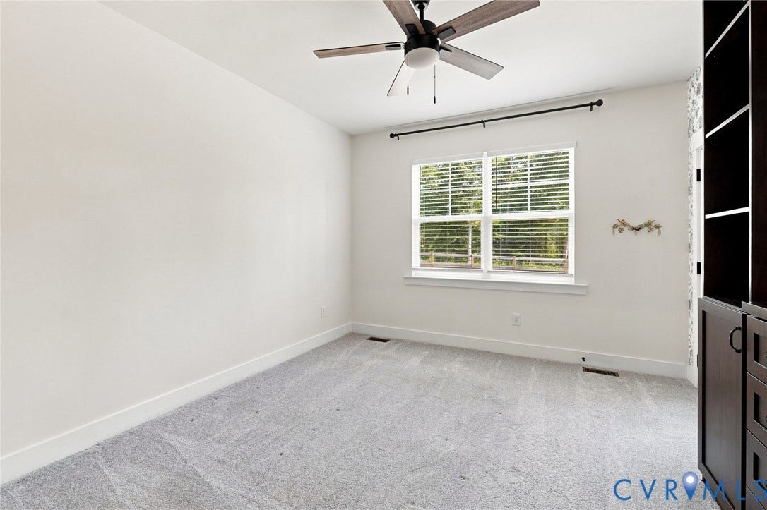 Light gray carpeting, a ceiling fan with light fixture, a window with horizontal blinds, and built-in cabinetry with shelving